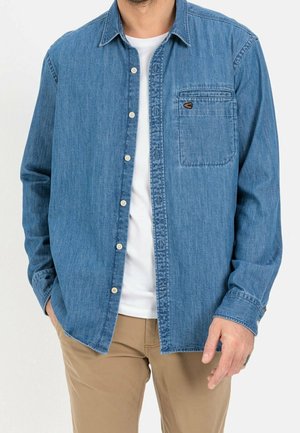 Mężczyzna w rozpiętej niebieskiej dżinsowej koszuli na białym t-shircie i beżowych spodniach, z rękami w kieszeniach na jednolitym tle.