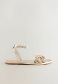 Sandale plate dorée métallique avec boucle à la cheville et bride avant tressée, présentée sur un fond clair uni.