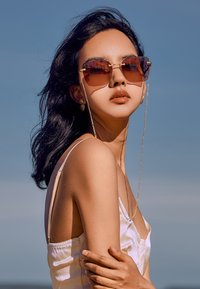 Sluneční brýle s oversized hnědými skly a zlatými detaily. Rám s texturou a řetízkovým akcentem. Model má na sobě světle zbarvený top.