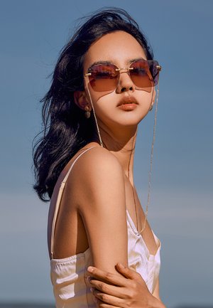 Occhiali da sole con lenti oversize di colore marrone e dettagli in tono oro. Montatura testurizzata con un accento a catena. Il modello indossa una maglietta di colore chiaro.