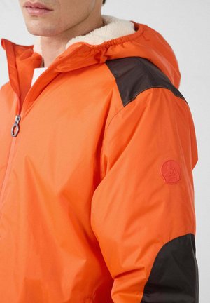 Chaqueta naranja con acentos negros, que cuenta con cierre frontal con cremallera, capucha con forro suave y texturizado, y un parche con logo en la manga.