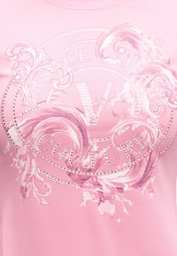 Pink fabric with a Versace Jeans Couture logo featuring a large "V," surrounded by decorative swirls and embedded rhinestones.