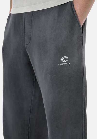 Pantalon gris décontracté avec poches latérales, porté par une personne avec la main dans la poche, arborant un logo blanc CamperLab sur la partie supérieure de la cuisse.