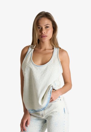 Donna con capelli castano chiaro indossa un top bianco senza maniche e dai tessuto strutturato e pantaloni abbinati, posando davanti a uno sfondo chiaro e uniforme.