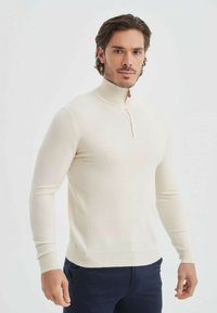 Un pull à col montant de couleur crème fabriqué en matériau à mailles fines. Il dispose d'une demi-fermeture éclair avec une tirette en cuir beige. Sa coupe est ajustée avec des manches longues.