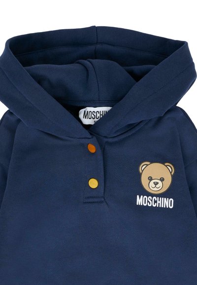 Felpa blu navale in morbido tessuto con grafica di un orso e testo "Moschino". Include tre bottoni a contrasto sul colletto.