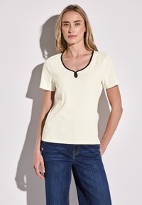 T-shirt a coste color crema chiaro con colletto rotondo bordato di nero e dettaglio con apertura a chiave sullo scollo. Maniche corte e design aderente.