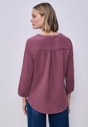 Nainen sivulta kuvattuna, yllään liilan sävyinen, teksturoitu pitkähihainen pusero, jossa on kerätty selän sauma, sekä siniset farkut, yksiväristä taustaa vasten.