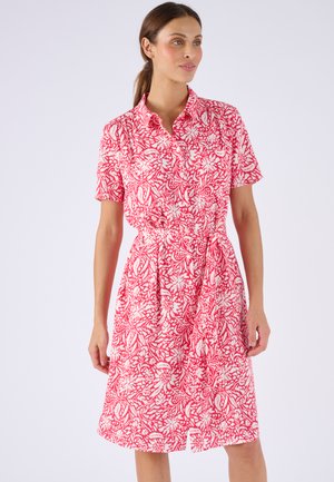 Mujer con vestido hasta la rodilla, de manga corta y cuello, con un estampado floral rojo y blanco y un cinturón atado en la cintura.