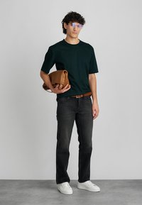 Cămașă scurtă, verde închis, blugi negri, adidași albi, ținând o geantă din piele maro. Design simplu, croială lejeră, stil casual.