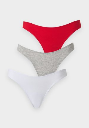 Junge Frau steht barfuß in einem weißen Crop-Top und roten Bikinihöschen vor einem einfachen weißen Hintergrund.
