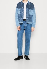 Kurtka dżinsowa z gradientem od jasnoniebieskiego do ciemnoniebieskiego, ozdobiona wzorami z logo. W zestawie z jasnoniebieskimi dżinsami i czarnymi butami na sznurowadła.