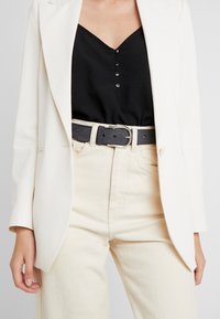 White blazer over a black V-neck top with button details, paired with high-waisted beige pants and a black belt with a metal buckle.