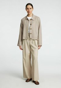 Chaqueta corta beige con botones, combinada con pantalones de pierna ancha beige claros. El modelo lleva una camisa blanca y zapatos planos trenzados marrones.