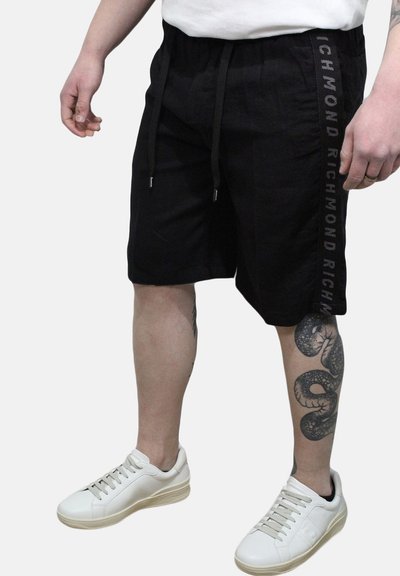 Pantaloni corti neri con cordino laterale, caratterizzati da una striscia verticale con logo. Abbinati a sneakers bianche, con un tatuaggio a forma di serpente dall'aspetto texturizzato sulla gamba.