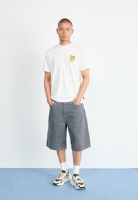 White cotton t-shirt with colorful fruit graphic on left chest, paired with gray denim shorts and gray and brown athletic shoes.