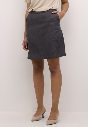 Kobieta ubrana w spódnicę do kolan w kolorze grafitowym, beżową bluzkę z krótkim rękawem oraz beżowe szpilki z ostrym noskiem, stojąca na tle jednolitego tła.