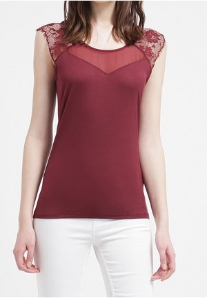 Vrouw draagt een mouwloos bordeauxrood topje met doorschijnende stof en kanten details op de schouders, gecombineerd met een witte broek.