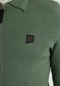 Pull zippé texturé vert avec une petite pièce noire présentant un design abstrait orange minimaliste sur la poitrine.