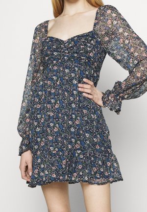 Kukallinen mekko, jossa on tummansininen tausta ja vaaleanpunaisia sekä valkoisia kukkia, muotoiltu bodice, neliön muotoinen pääntie ja pitkät läpikuultavat hihat.