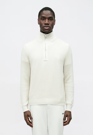 Weißer, gerippter Reißverschluss-Pullover mit hohem Kragen, ausgestattet mit einem halben Reißverschluss und einem strukturierten Strickmuster, kombiniert mit hellen Hosen.