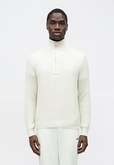 Pull blanc côtelé à fermeture éclair avec un col montant, doté d'une demi-fermeture éclair et d'un motif en maille texturée, associé à un pantalon de couleur claire.