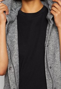 Szary dzianinowy kaptur z zamkiem błyskawicznym i zatrzaskami, o miękkiej fakturze. Pod spodem noszona jest czarna koszulka. Kaptur opada na ramiona.