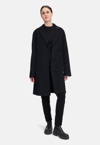 Sort uldfrakke med klippet krave, dobbelt knappelukning og oversized pasform. Båret over en sort bluse og smalle sorte bukser med kraftige hæl støvler.