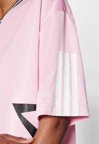 Camiseta deportiva rosa con un triángulo gráfico negro y la marca de tres rayas blanca en la manga, fabricada con una textura de tela suave y transpirable.