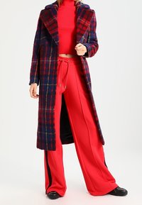 Abrigo de cuadros largo en azul marino, rojo y verde con un cuello ancho, combinado con pantalones de chándal rojos de cintura alta con franjas laterales negras.