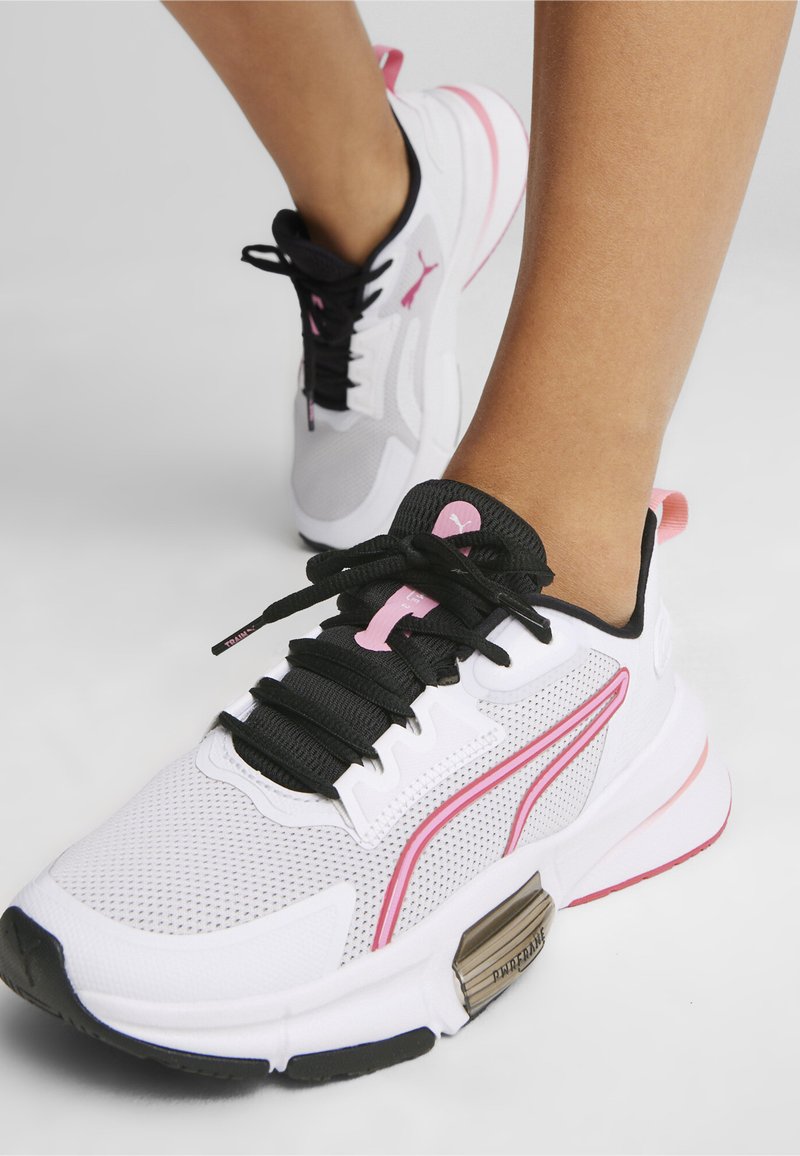 Zapatillas deportivas blancas con parte superior de malla, cordones negros, acentos rosas y una suela texturizada. Presenta un detalle de marca prominente en el costado.