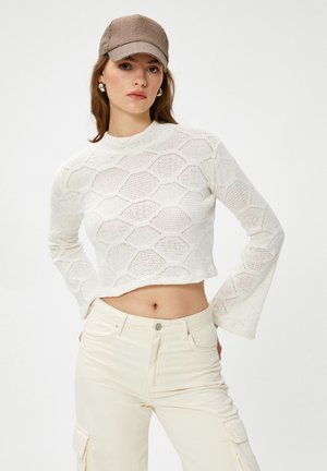 Femme portant un pull en tricot crémeux court avec des manches évasées, un pantalon cargo ivoire, des boucles d'oreilles en argent et une casquette à carreaux marron.