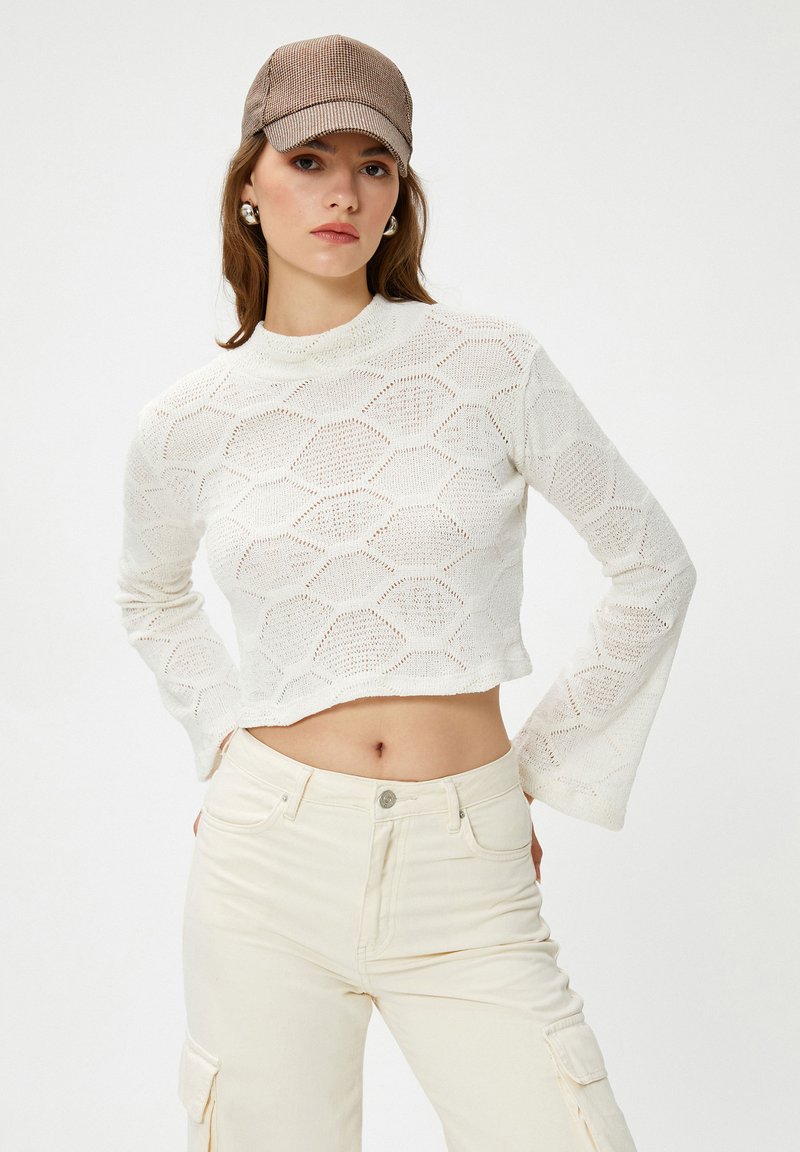 Frau in einem cremefarbenen gestrickten cropped Pullover mit Glockenärmeln, elfenbeinfarbenen Cargo-Hosen, silbernen Creolen und einer braunen karierten Mütze.