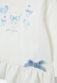 Haut en coton blanc avec un imprimé ourson et des noeuds bleus. Ourlet volanté orné d'un petit noeud en velours bleu sur le côté droit.