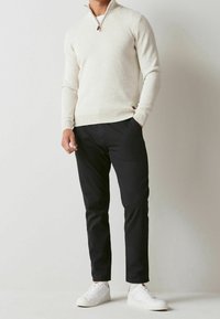 Light beige zip-neck sweater made of soft material, paired with black pants and white sneakers, showcasing a casual outfit.
