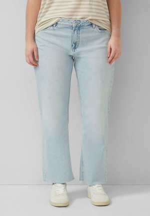 Osoba ubrana w jasno-niebieskie dżinsy o prostych nogawkach, beżową koszulę w paski oraz białe trampki, stojąca na tle jednolitego szarego tła.