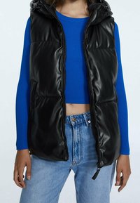 Chaleco acolchado negro con un acabado brillante, cierre de cremallera y capucha, llevado sobre una blusa de punto azul. Combinado con jeans de mezclilla azul claro.