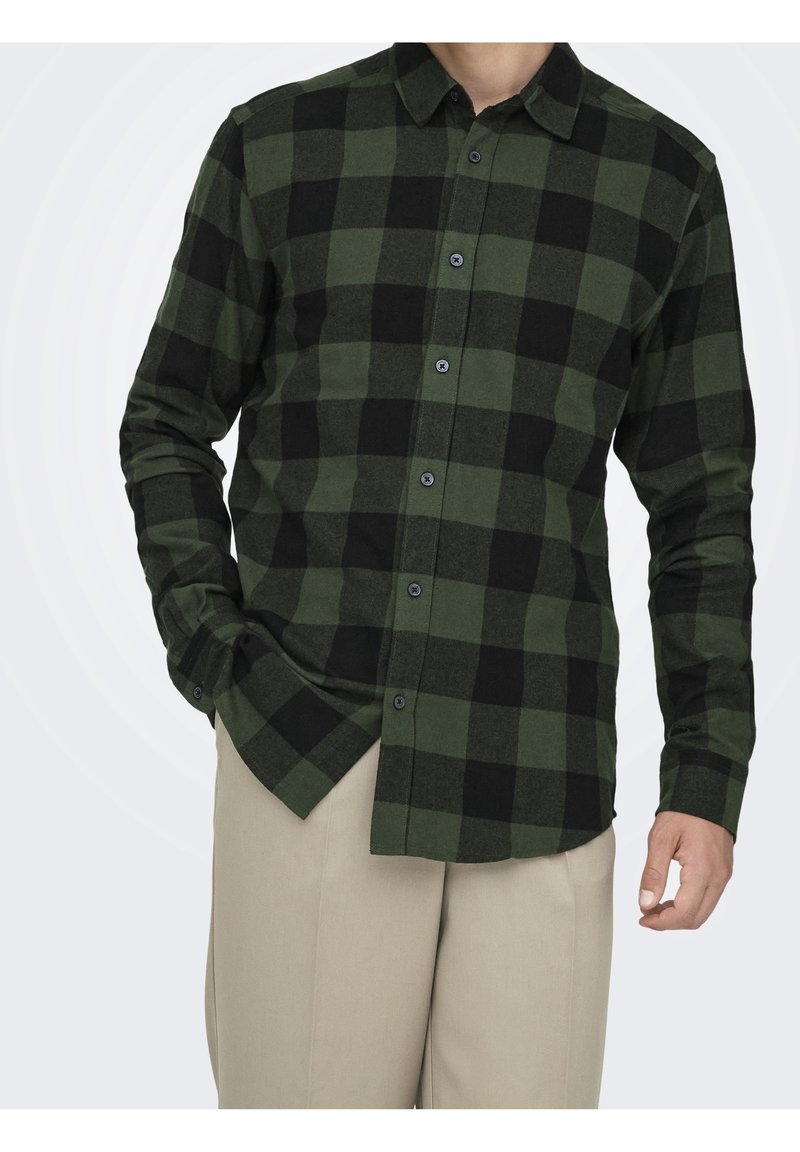 Homme portant une chemise à carreaux verts et noirs à manches longues boutonnée avec un pantalon beige, main partiellement dans la poche.