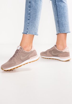 Person, der bærer lysebrune Nike-sneakers med hvide mellemsåler og gummisåler, kombineret med lyseblå croppede jeans, står på en hvid gulv.