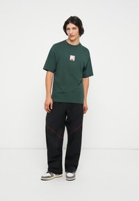 Camiseta de algodón verde con mangas cortas y un logo cuadrado. Pantalones negros con ribetes rojos y corte relajado. Zapatillas blancas y grises.