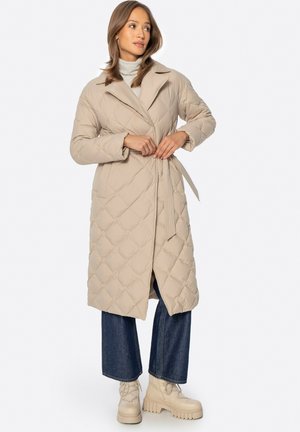 Vrouw die een beige doorgestikte lange jas met riem dichtmaakt, draagt een grijze coltrui, wijde jeans en stevige beige laarzen, staand op een witte achtergrond.