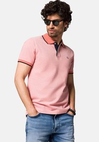 Jasnoróżowa koszulka polo z koralowym kołnierzem i granatowymi akcentami, krótki rękaw oraz małe logo. Nosi z niebieskimi dżinsami i czarnymi okularami przeciwsłonecznymi.