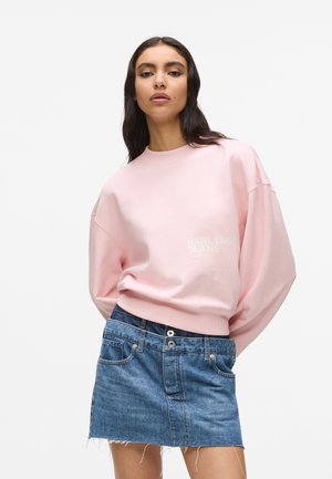 Žena v růžovém oversized svetru s nápisem "KARL LAGERFELD JEANS" a modré džínové mini sukni na jednobarevném bílém pozadí.