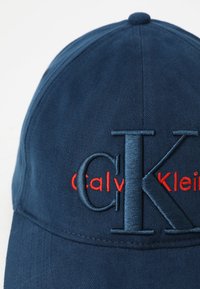 Blaues Baseballcap mit einem erhabenen "CK"-Logo in Dunkelblau und "Calvin Klein" in roter Stickerei. Der Stoff hat eine strukturierte Oberfläche.