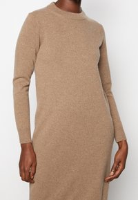 Robe en tricot beige avec un col ras du cou côtelé et des manches longues, présentant une texture douce et une coupe ajustée qui épouse délicatement la silhouette.