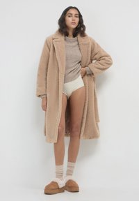Donna che indossa un lungo cappotto teddy beige, un maglione taupe, biancheria intima crema, calze beige e pantofole con pelliccia tan, in piedi su uno sfondo bianco.