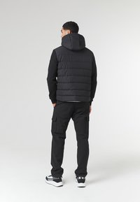 Sort puffer vest med hætte, prydet med quiltet design, båret over en sort langærmet skjorte, parret med sorte cargo-bukser og sneakers.