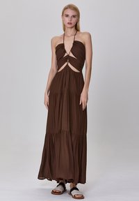 Robe maxi marron avec un col halter, des découpes, et une jupe tiered. Fabriquée en tissu léger, elle présente une silhouette fluide et un minimum de métal.