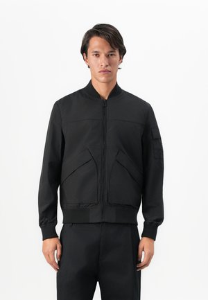 Black bomber jacket made of smooth fabric. Features a front zipper, ribbed cuffs, and two angled side pockets. Minimalist design.