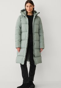 Polstret lang jakke i lys grøn, der har hætte, trykknapper og en glat tekstur. Bæres over en sort top og mørke jeans.